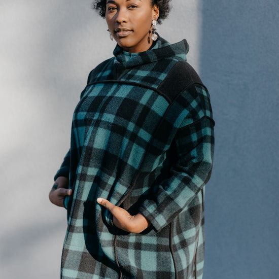 Black woman standing outside in front of a grey wall wearing a green and black plaid siberian parka.  Her hands are in the pockets.