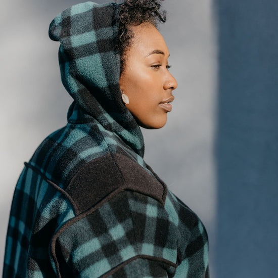 Close up of Black woman standing outside in front of a grey wall wearing a green and black plaid siberian parka. The hood is up and she is facing to the right.