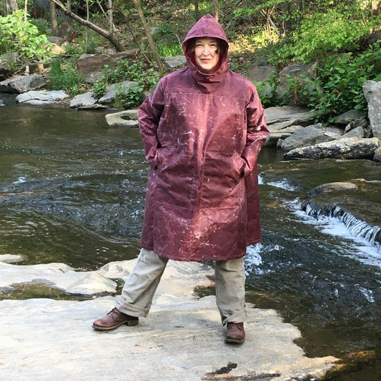 Woman standing on a rock in a creek wearing the parka made of maroon waxed cotton . Her hands are in her pockets.