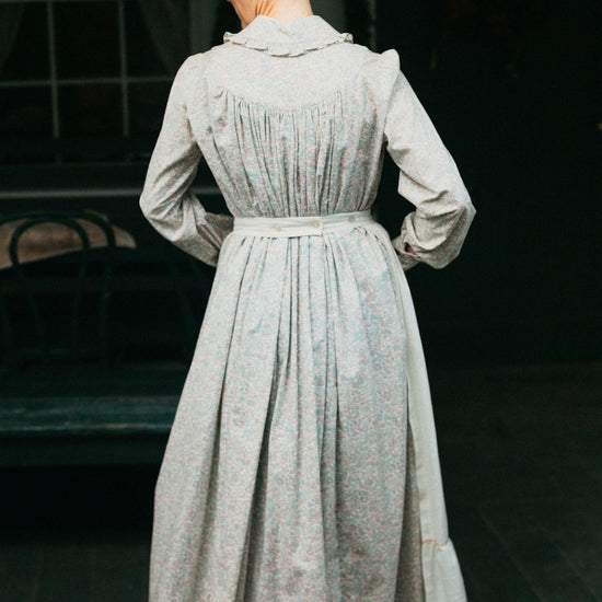 Photo of back view of prairie dress and apron waistband.  Model stands outside with hands in front pocket.