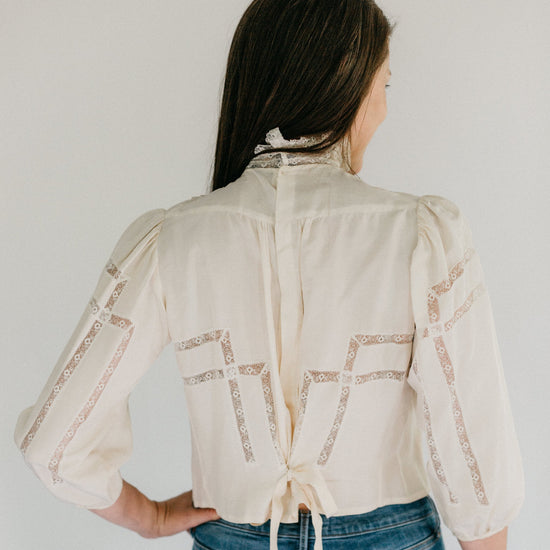 Back view of brunette woman standing in front of a white studio backdrop, wearing 205 Gibson Girl Blouse with hand left hand on her hip.