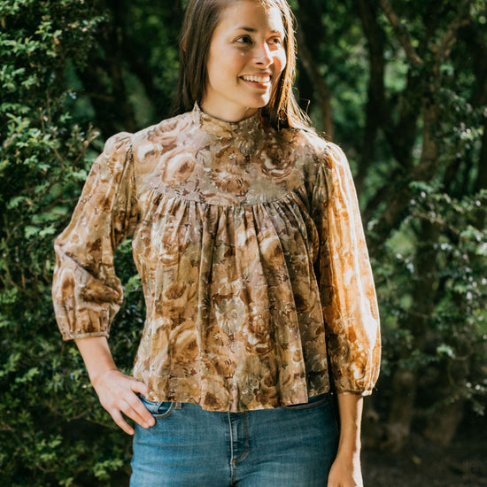 Brunette woman standing surrounded by greenery with hand on her hip looking to the right, wearing 205 Gibson Girl Blouse.