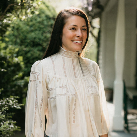 Brunette woman smiling looking to the right,  surrounded by greenery wearing 205 Gibson Girl Blouse.