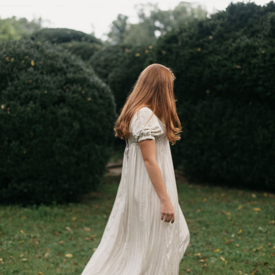Side view of red head young woman standing surrounded greenery wearing 215 Empire Dress.