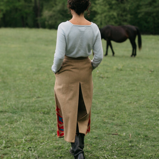 Back view of woman walking surrounded bu greenery with hands in the pocket of the culotte 231 Big Sky Riding Skirt, with a horse in the background.
