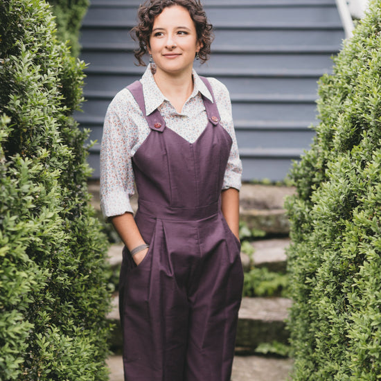 Young brunette white woman standing with hands in pockets of the 240 Rosie the Riveter overalls surrounded by greenery.