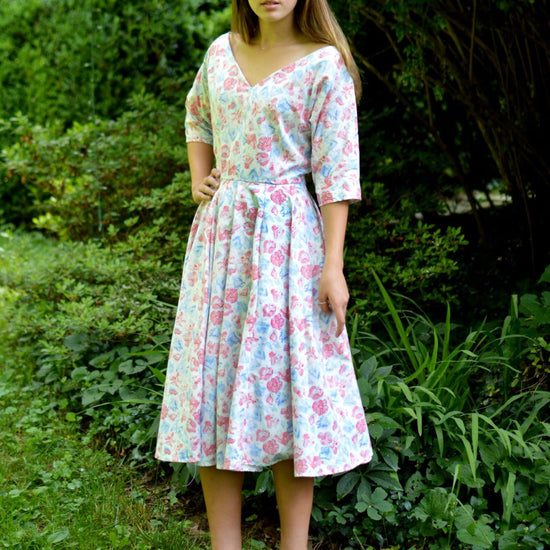 Young woman standing in front of trees and shrubs wearing a white and pink floral print dress with a v-neck.