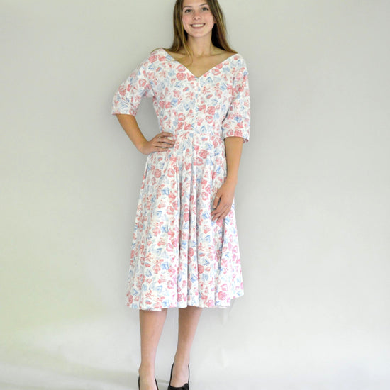Young woman smiling wearing a below-knee length white and pink floral print dress with a v-neck and 3/4 length sleeves and black shoes.  She is in front of a white background.
