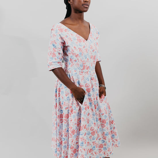 Young woman wearing a below-knee length white and pink floral print dress with a v-neck and 3/4 length sleeves and black shoes.  She is in front of a white background and her hands are in the skirt pockets.