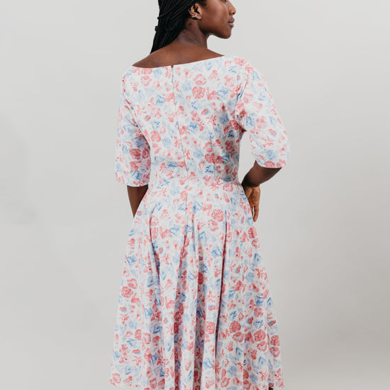 Young woman with her back to viewer is wearing a below-knee length white and pink floral print dress with a v-neck and 3/4 length sleeves and black shoes.  She is in front of a white background.