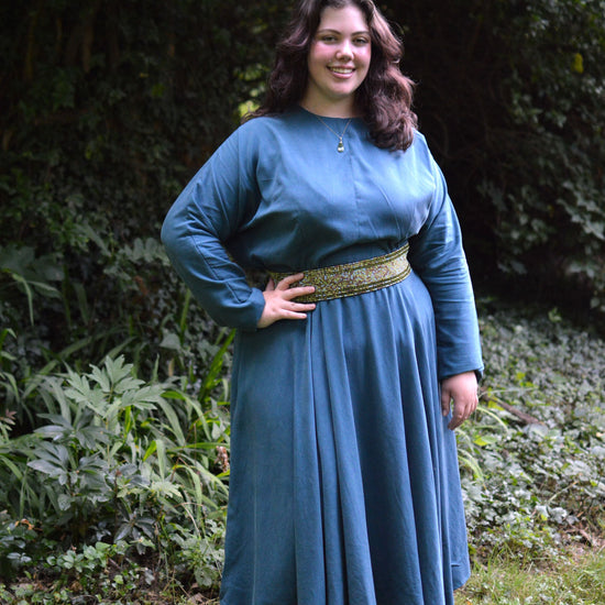 Young woman wearing a blue long sleeve, jewel neck, calf-length dress outside in front of shrubbery.  She has on a green a white beaded belt and black shoes. Her right hand is on her hip.