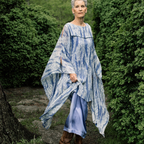 Older white woman with short grey hair standing surrounded by greenery,wearing brown boots and the Greek Island Dress Tunic displaying the flare of the free- floating sleeve panels.