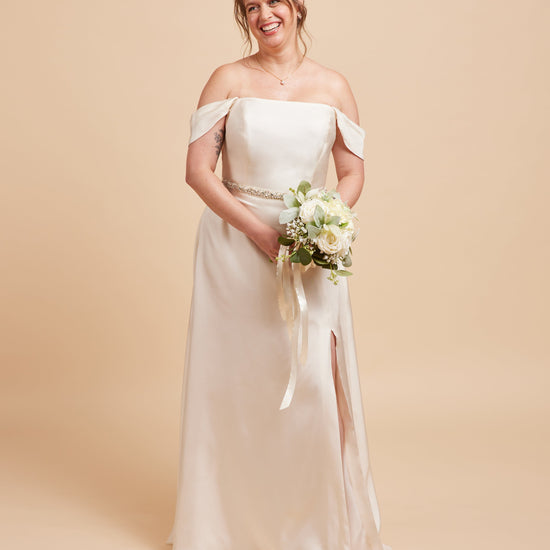 Woman in a white off-shoulder wedding dress holding a bouquet on a beige background. This dress is the Ellington Strapless Dress sewing pattern from Cashmerette.