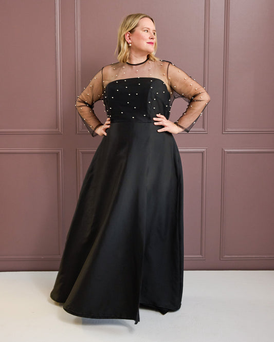 Woman wearing a black evening gown with sheer sleeves against a pink paneled wall. This is the Ellington Dress sewing pattern from Cashmerette.