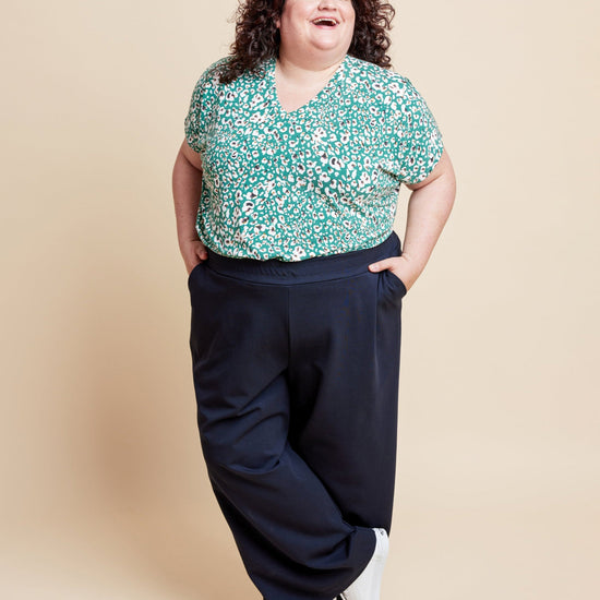 Woman wearing a floral top and dark pants on a beige background. This is the Grafton Dress, Top & Skirt sewing pattern from Cashmerette.