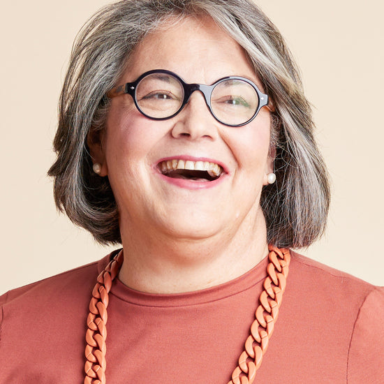 Person with gray hair wearing glasses and a brown shirt with a large orange necklace on a beige background. This is the Grafton Dress, Top & Skirt sewing pattern from Cashmerette.