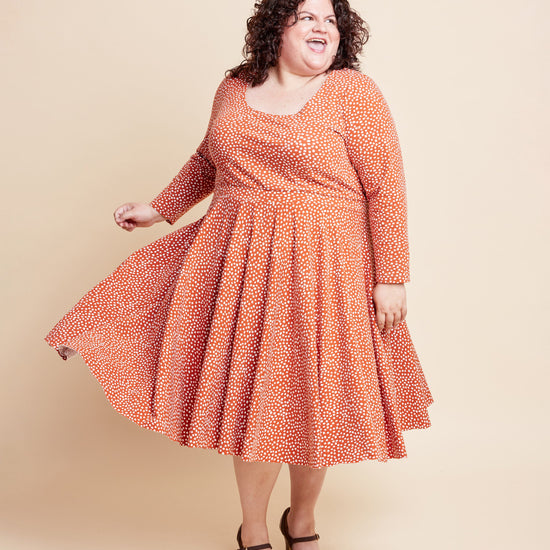 Woman wearing an orange patterned dress against a beige background. This is the Grafton Dress, Top & Skirt sewing pattern from Cashmerette.