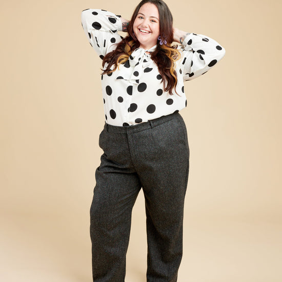 Person wearing a polka dot blouse and gray pants on a beige background. These pants are the Meriam Trousers sewing pattern from Cashmerette.