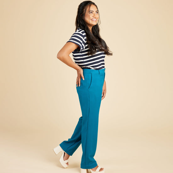 Woman wearing a black and white striped shirt and blue pants on a beige background. These pants are the Meriam Trousers sewing pattern from Cashmerette.
