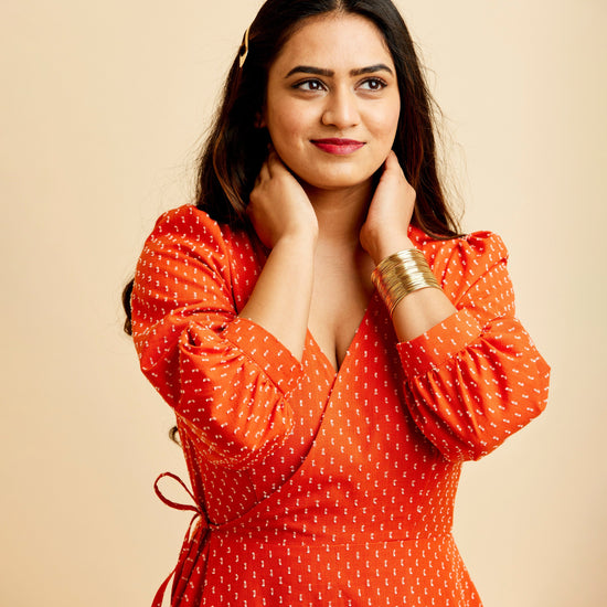 Woman wearing an orange dress with a beige background. This dress is the Roseclair Wrap Dress sewing pattern from Cashmerette.