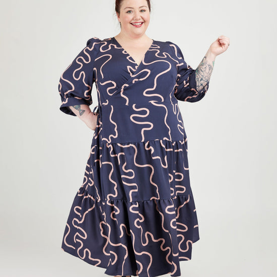 Woman wearing a navy dress with pink wavy patterns on a white background. This dress is the Roseclair Wrap Dress sewing pattern from Cashmerette.