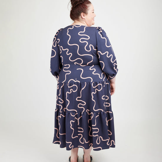 Woman wearing a navy dress with pink wavy patterns on a white background. This dress is the Roseclair Wrap Dress sewing pattern from Cashmerette.