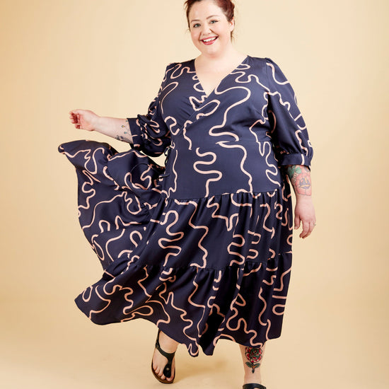 Woman wearing a navy dress with wavy pattern on a beige background. This dress is the Roseclair Wrap Dress sewing pattern from Cashmerette.