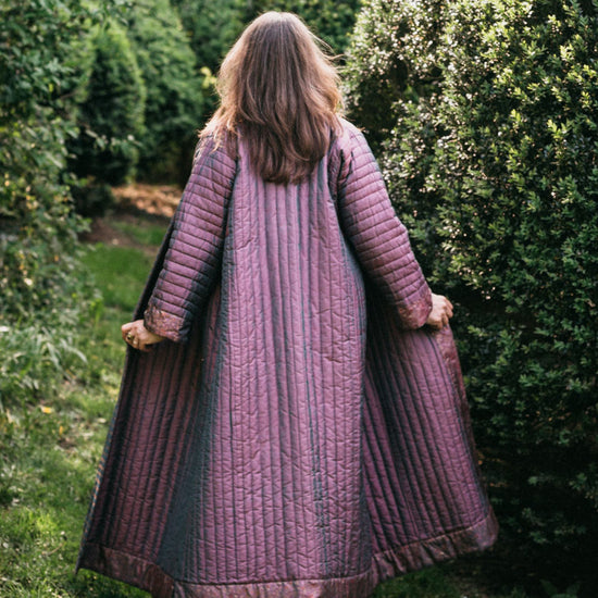 Back view of model walking away from camera outdoors wearing lavender silk Turkish coat.  Coat has full trapunto quilting and is ankle length. Model is holding jacket out at hip level showing full width.