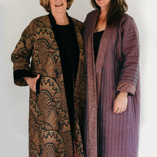 Photograph of two women standing side by side.  Woman on the left is wearing a tapestry fabric Turkish coat with trapunto trim around sleeves, front opening and bottom hem. Woman on right is wearing a solid lavandar silk coat with trapunto stitching all over the body of coat.