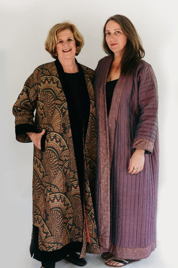 Photograph of two women standing side by side.  Woman on the left is wearing a tapestry fabric Turkish coat with trapunto trim around sleeves, front opening and bottom hem. Woman on right is wearing a solid lavandar silk coat with trapunto stitching all over the body of coat.