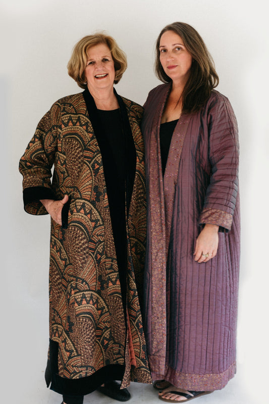 Photograph of two women standing side by side.  Woman on the left is wearing a tapestry fabric Turkish coat with trapunto trim around sleeves, front opening and bottom hem. Woman on right is wearing a solid lavandar silk coat with trapunto stitching all over the body of coat.